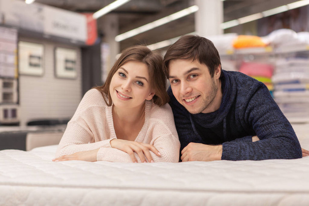 Attractive young woman and her cheerful handsome boyfriend embracing, lying on the bed together at home furnishings store. Happy couple buying orthopedic mattress and bed for their new home - Fotografie, Obrázek