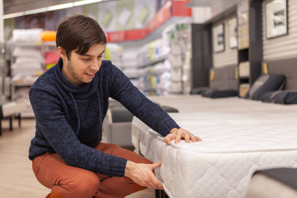 Handsome cheerful man examining orthopedic mattress at furniture store, copy space. Young man shopping at home furnishings department, choosing new bed to buy. Rest, relaxation, health concept - Фото, изображение