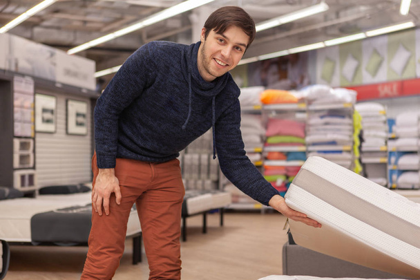 Attractive happy man examining mattress on sale, shopping at home goods supermarket department. Young male customer choosing new bed to buy. Lifestyle, living, comfort concept - Photo, Image