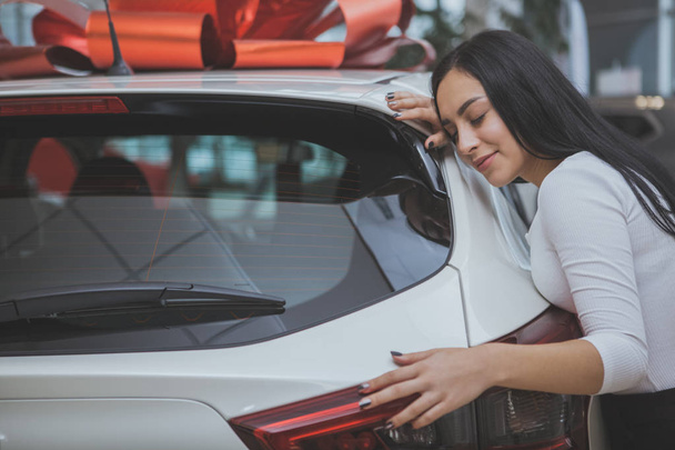Charming young woman smiling with her eyes closed, hugging her newly bought automobile at the dealership. Happy woman embracing her new car with a red bow on the roof. Car present concept - Photo, image
