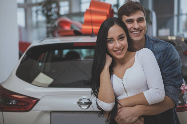 Gorgeous young happy couple hugging, smiling to the camera, standing in front of their new car, copy space. Handsome man embracing his lovely girlfriend after buying new automobile. Family car concept - Φωτογραφία, εικόνα