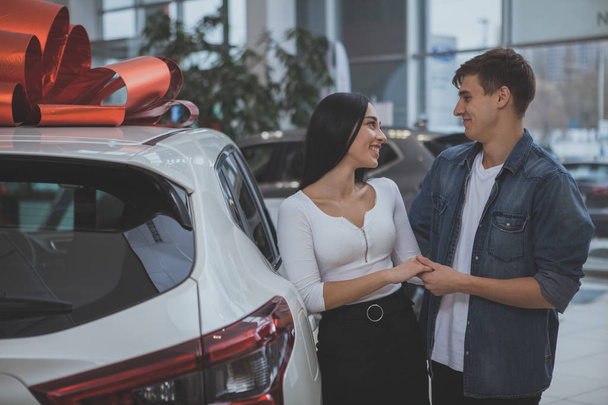 Happy young couple holding hands smiling at each other while choosing new car at the dealership salon. Cheerful beautful woman and her husband buying new auto. Transportation vehicle concept - Valokuva, kuva