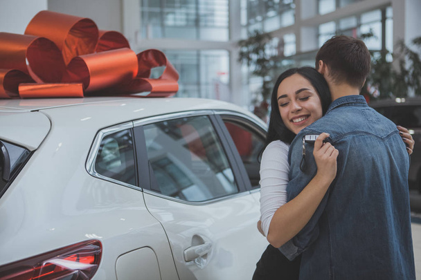 Gorgeous woman hugging her husband, standing near new car with red bow on the roof. Loving man buying new automobile to his beautiful wife, copy space. Couple hugging, celebrating new auto - 写真・画像
