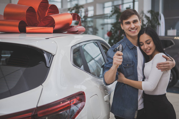 Handsome happy man smiling joyfully, hugging his gorgeous girlfriend after buying new car at the dealership. Lovely couple holding car keys, posing near their new automobile with red bow on the roof - Foto, immagini