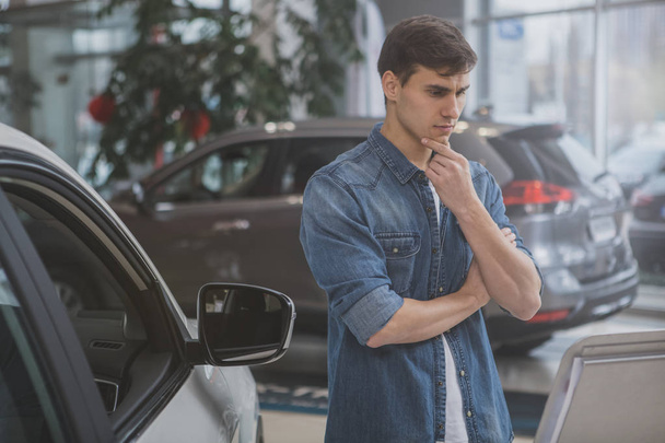 Young attractive man looking at new car far sale thoughtfully, rubbing his chin. Handsome male customer choosing new automobile at dealership salon. Thoughtful man shopping for a new car, copy space - Photo, Image