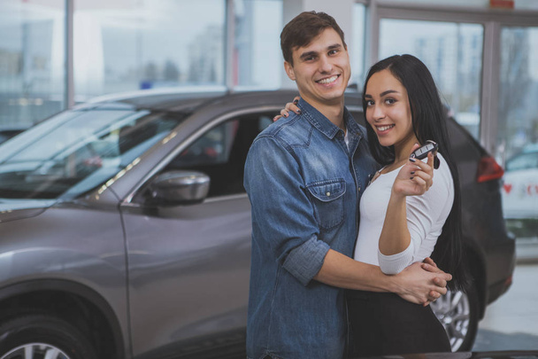 Handsome happy man embracing his gorgeous girlfriend in front of their new automobile at the dealership, copy space. Beautiful woman smiling, holding car keys, after buying new auto with her husband - Fotografie, Obrázek