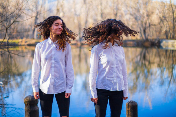 Young twin sisters in same clothes shaking hair at lake on autumn day in the forest - Photo, Image