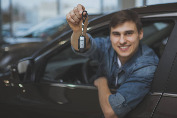 Cheerful male driver holding out car key to the camera, smiling joyfully after buying new automobile. Happy young man sitting in his new auto at the dealership salon. Owner, rental service concept - Foto, afbeelding