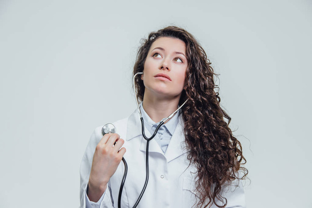 A serious young woman in a Caucasian woman in a laboratory robe, looking at you and holding the phonendoscope in his hands has a surprised look. Portrait of a woman is pretty dark hair - a doctor - 写真・画像