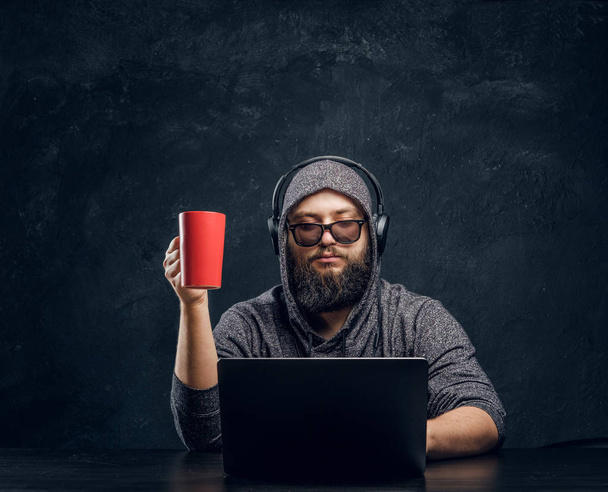 Un hacker se sienta detrás de un portátil, sosteniendo una taza de café y trabajando con programas en una habitación oscura
 - Foto, imagen