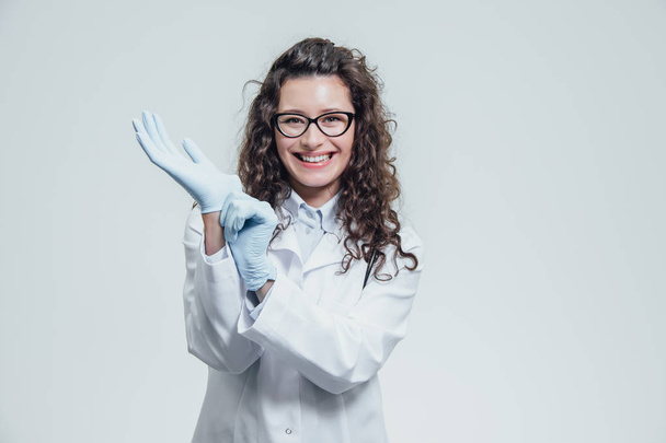 Young woman doctor wearing glasses in blue gloves looks intently into the camera. Isolated on a gray background - Photo, image