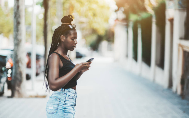 A cute young black girl with braided hair is standing on a bright city street is checking her route on the map via the smartphone; a charming African teenage African female typing message on cellphone - Valokuva, kuva