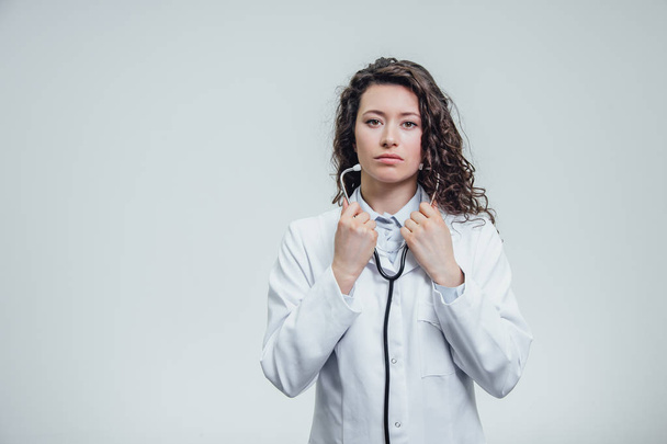 A serious young female Caucasian woman in a laboratory robe, looking at you and holding the phonendoscope out of hand. Portrait of a pretty dark hair woman - a doctor wearing white uniforms. The - Foto, Bild