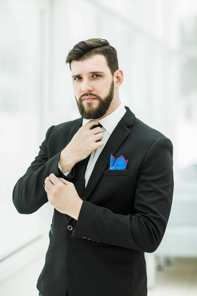 successful businessman adjusting his necktie while standing near a window in bright office. - Foto, Bild