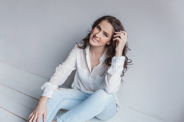 Happy teenage girl smiling. Young happy positive woman in white shirt sitting on floor in bright room against white wall. European woman. Positive human emotion facial expression body language - Foto, Imagen
