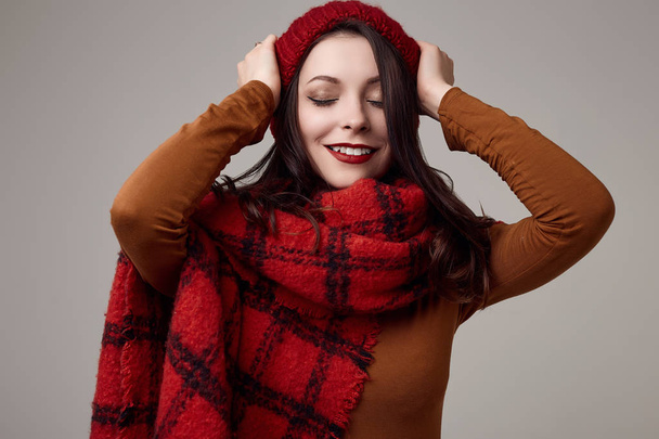 Fashion portrait of beautiful brunette woman in sweater and red hat with big knitted scarf on white background in studio - Photo, Image