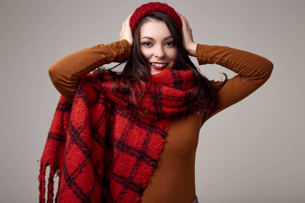Fashion portrait of beautiful brunette woman in sweater and red hat with big knitted scarf on white background in studio - Photo, Image