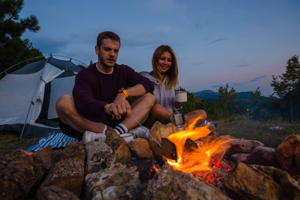 Young couple drinking coffee and warming up on the campfire in the forest hill in the dusk - Photo, Image