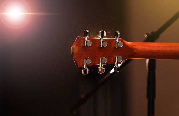 Akoestische gitaar hoofd met pinnen, nek, frets en toets, achteraanzicht tuning. Kop van de gitaar over concert podium achtergrond met licht flare effect. - Foto, afbeelding
