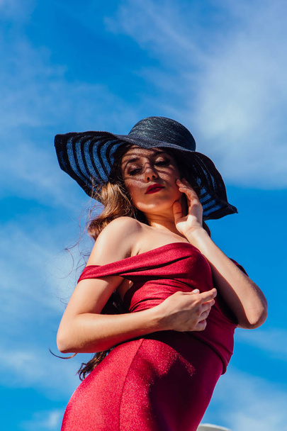 Beautiful young woman in red dress and balack hat in summer sunny day on the blue sky background. Retro fashion - Foto, afbeelding