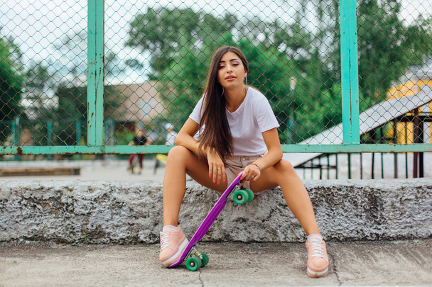 Summer lifestyle image of trendy pretty young girl sitting next to the skateboard coart with her purple plastic skateboard. - Photo, Image