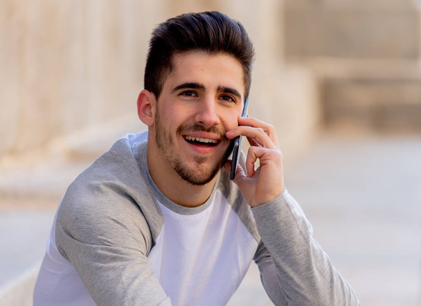 Young attractive college man in his twenties happy talking and chatting with friend on smart mobile phone on campus or city stairs. In communication, technology internet and travel around europe. - Foto, immagini