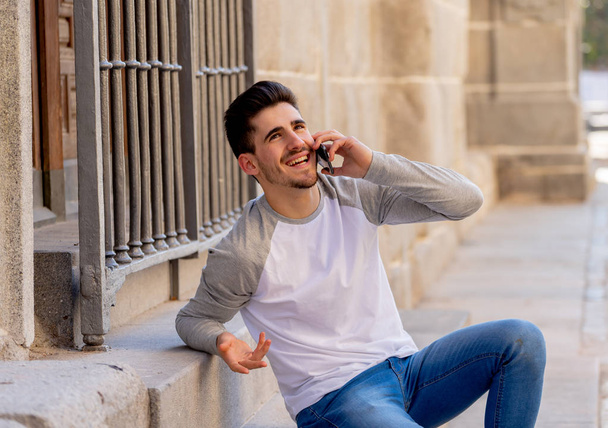 Young attractive college man in his twenties happy talking and chatting with friend on smart mobile phone on campus or city stairs. In communication, technology internet and travel around europe. - Фото, изображение