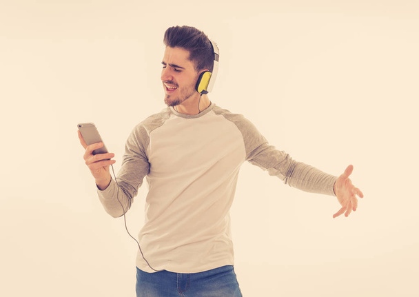 Portrait of happy young man listening to music in headphones changing songs and tracks on smartphone and singing and dancing. Isolated white background. In positive emotions, youth happiness concept. - 写真・画像