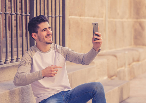 Excited trendy young man taking a selfie on smart mobile phone on holidays in european city street or in university campus outside. In holidays and technology, connection and social media concept. - Foto, Imagem