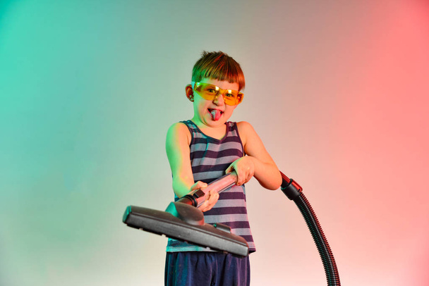 Ένας μικρός βοηθός με γυαλιά, κατευθύνει τη βούρτσα της ηλεκτρικής σκούπας προς τα εμπρός, φωτογραφία στο στούντιο σε ένα κόκκινο-μπλε φόντο. - Φωτογραφία, εικόνα