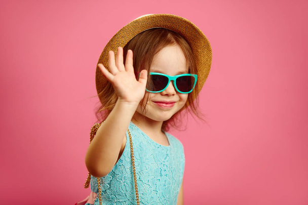 Little cute girl in summer hat and sunglasses waving at you,expressing joy and happiness, standing on pink isolated background. - Фото, изображение