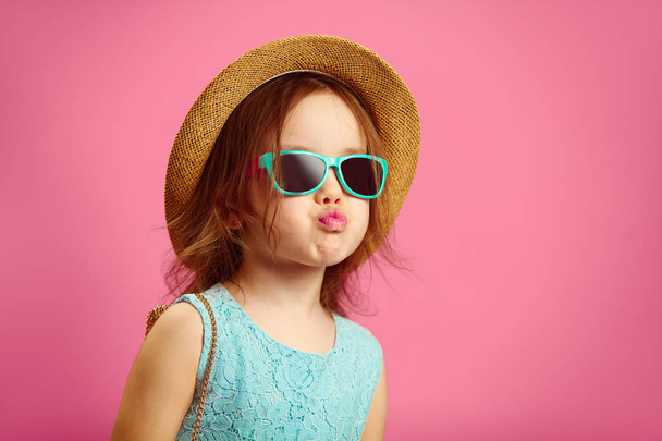 Beautiful little girl dressed in fashionable clothes, wears straw hat, sunglasses and blue dress, shows duck face, stands on pink isolated. - Foto, Bild