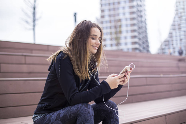 Молодая спортивная женщина отдыхает и слушает музыку на мобильном телефоне после тренировки на открытом воздухе в городской среде
 - Фото, изображение