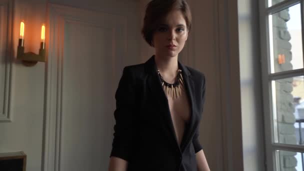 A woman with a short haircut, dressed in a black jacket on a naked body goes to the camera, holds a glass of champagne in her hands, stops and drinks champagne, then goes back to the camera and stops  - Video, Çekim