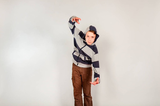 The boy in the hood holds his hands up on a gray background in the studio - Photo, Image