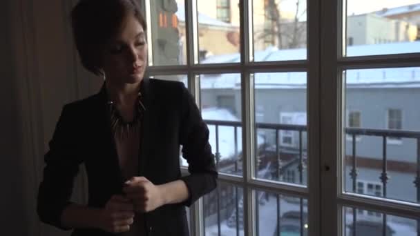 A woman with a short haircut and brown hair in a black pantsuit, with a jacket on her naked body standing at the big window, posing for the camera, looking arrogant, then smiling, spinning and posing  - Filmagem, Vídeo