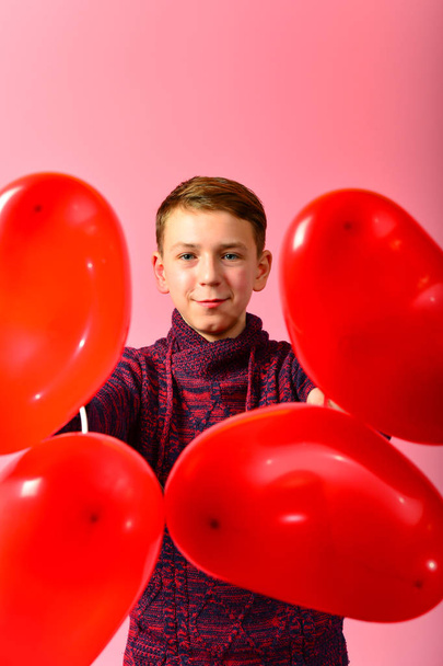 Мальчик среди красных сердечных шариков в виде сердечка на розовом фоне на День Святого Валентина и 8 марта
. - Фото, изображение