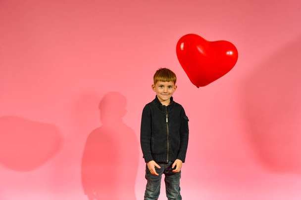 Ένα αγόρι πετάει και πιάνει ένα μπαλόνι σε σχήμα καρδιάς σε ένα ροζ φόντο στο στούντιο. - Φωτογραφία, εικόνα