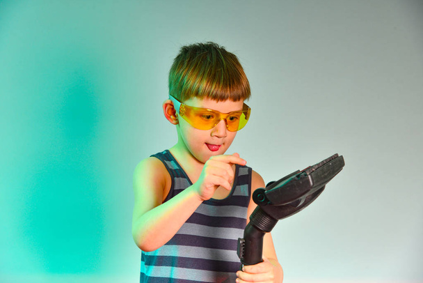 Το αγόρι με τα κίτρινα γυαλιά καθαρίζει τη βούρτσα της ηλεκτρικής σκούπας, φωτογραφία στο στούντιο. - Φωτογραφία, εικόνα