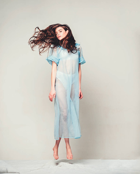 Lady in transparent blue nightie and white lingerie fly above bed, grey background. Woman with long curly hair wears tender pajama. Girl on calm face jump on bed. Femininity concept. - Photo, Image