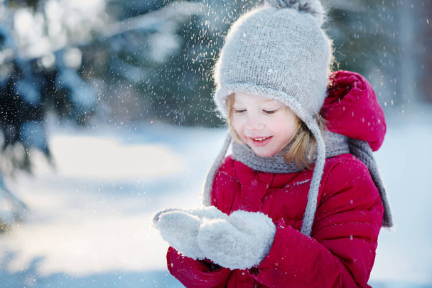  klein meisje in een rode jas zit in de sneeuw en sneeuwvlokken vangsten. Zonnige winterdag - Foto, afbeelding