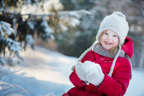μικρό κορίτσι σε ένα κόκκινο παλτό κάθεται στο χιόνι και χαμογελαστός. - Φωτογραφία, εικόνα