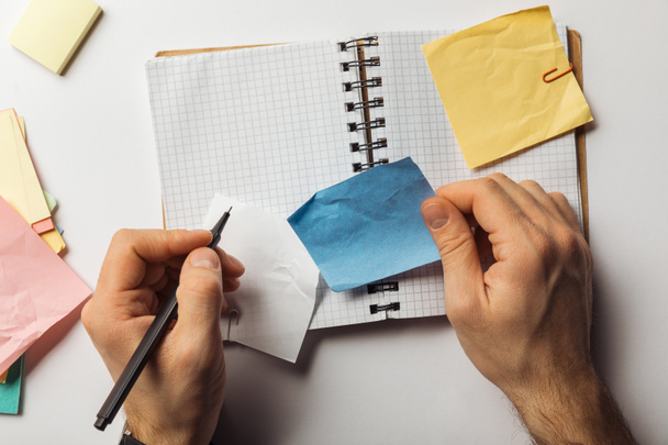 cropped view of man writing on sticky note and holding crumpled blue paper near opened notebook  - Photo, Image