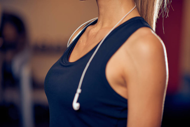 Close up of woman's breasts in sport top. Around neck earphones. Gym interior, healthy lifestyle concept. - Photo, Image