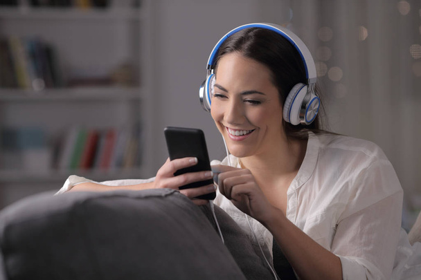 Happy girl listening to music checking phone sitting on a couch in the night at home - Fotografie, Obrázek