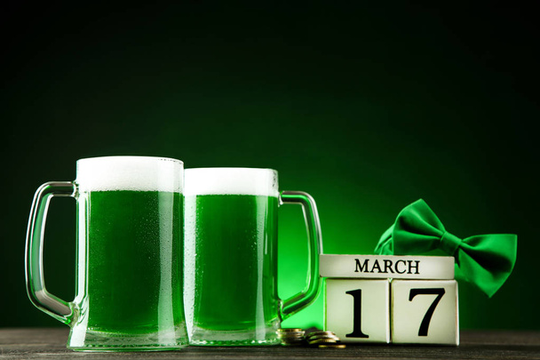 St. Patrick's Day. Green beer in mugs with cube calendar, coins and bow tie on dark background - Photo, image