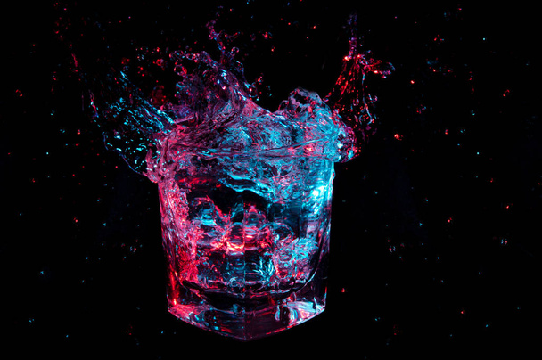 Огромный брызг прозрачной жидкости, светящейся синим и красным светом в скалах, стекло изолировано на черном фоне
 - Фото, изображение
