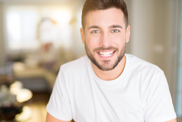 Jonge knappe man dragen casual wit t-shirt thuis met een gelukkig en cool glimlach op het gezicht. Gelukkig persoon. - Foto, afbeelding