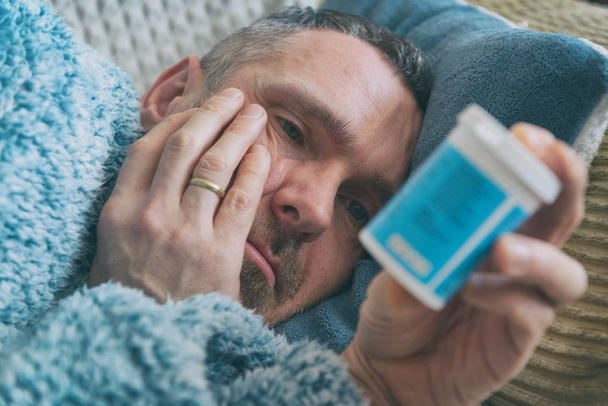 Зрілий чоловік, що страждає депресією, лежить сумно вкритий ковдрою на ліжку і тримає пляшку антидепресантів в руці
 - Фото, зображення
