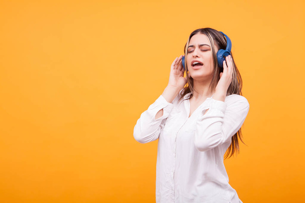 Portrait of excited girl singing and listening music on blue headsphones on yellow background - Фото, зображення